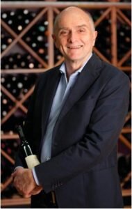 Profile photo of Peter Scudamore-Smith standing in front of a wine rack holding a bottle of wine.